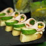 Elegant canapés with cucumber, ham, and garnish served on baguette slices with cocktails.